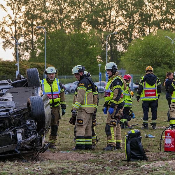 Räddningspersonal arbetar vid en bil som voltat och hamnat upp och ner på en gräsplan.