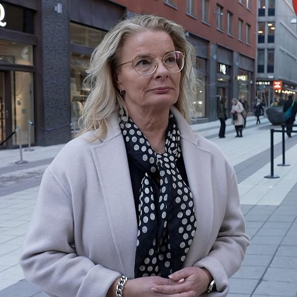 Kvinna klädd i ljus kappa och prickig halsduk står på en gågata i stadsmiljö, medan en mikrofon med ”SVT”-märke är riktad mot henne, vilket indikerar en pågående intervju.