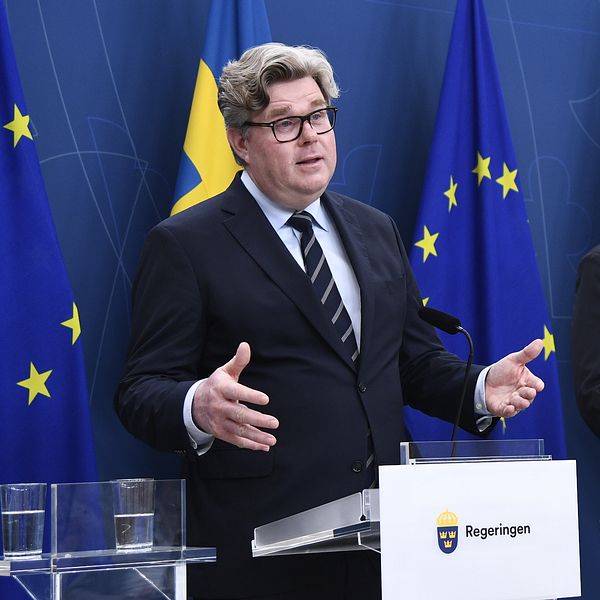 Två personer i kostym talar vid podier framför svenska och EU-flaggor på en presskonferens.