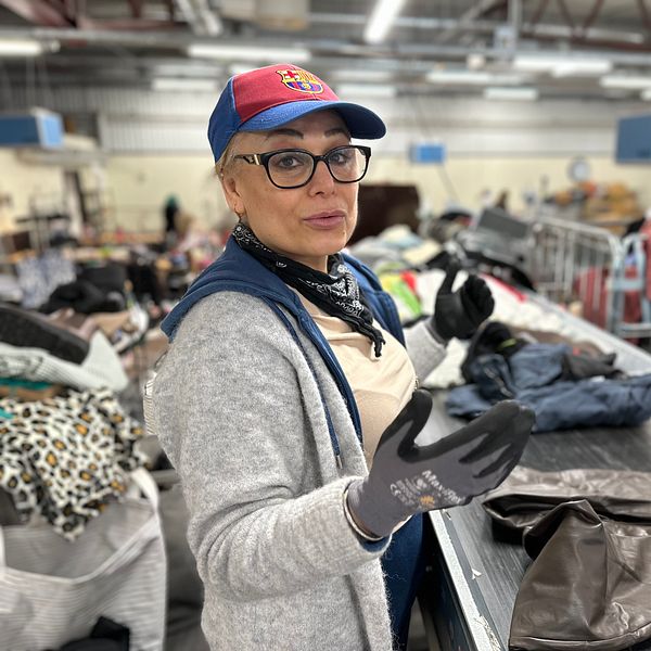 Madlin Benjamin står vid ett sorteringsband i en anläggning fylld med kläder och textilier. Personen bär en keps, handskar och en scarf samt en tröja. Runt omkring syns högar av kläder i olika färger och mönster, samt skyltar och infrastruktur från anläggningen.