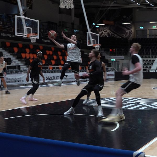Basketspelare tränar på plan i sporthall. Flera spelare in action vid korgen.