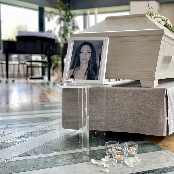 En kista placerad i ett ljust rum, omgiven av ett blommarrangemang och ljus. Framför kistan står en ram med ett fotografi, och på golvet ligger några tända ljus.