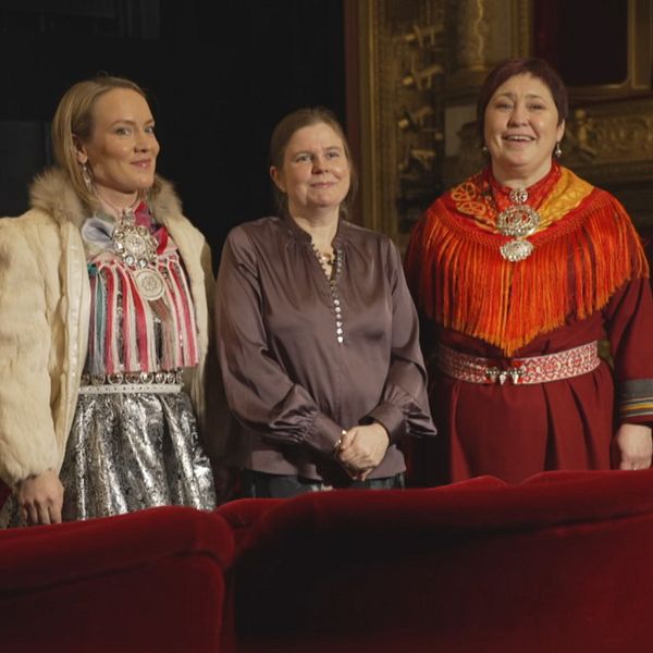 Tre personer iförda traditionella kläder står framför en ornant scen i en teatersalong. Till höger avbildas en del av en stor byggnad med en stenbågad entré och texten ”OPERAN” i guld.