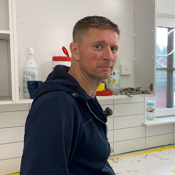 En person står vid en köksbänk i ett hem, omgiven av en arbetsyta med verktyg och material. Rummet har vita väggar och ett fönster som släpper in naturligt ljus. Det finns hyllor med rengöringsmedel och verktyg i bakgrunden.