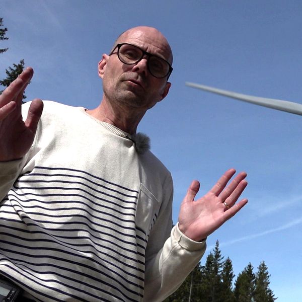 Person gestikulerar framför ett vindkraftverk mot blå himmel och trädtoppar.