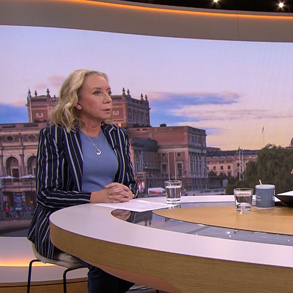 Tre personer sitter vid ett rundat bord i en TV-studio. Bakom dem syns en stadsskyline med byggnader och en färgglad himmel, vilket antyder att det är morgon eller sen eftermiddag. De två personerna som är vända mot kameran diskuterar medan den tredje personen, som sitter snett mot kameran, lyssnar. På bordet finns kaffemuggar och surfplattor.
