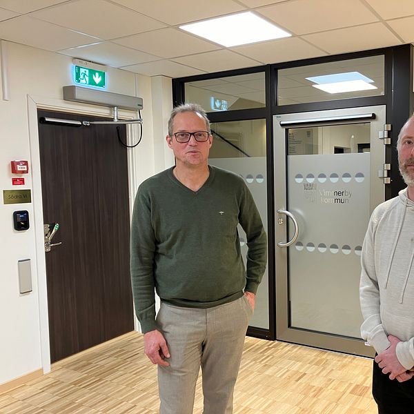 Två personer står i en korridor framför en glasdörr. Den första personen bär en grön tröja och ljusa byxor, medan den andra har en ljusgrå sweatshirt på sig. Korridoren har en modern inredning med trägolv och vita väggar. Bakom dem finns en dörr med dekaler som indikerar en kontorsmiljö.