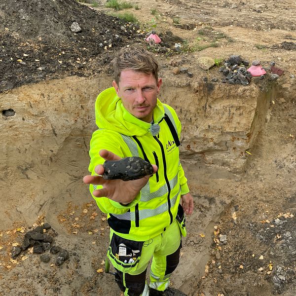 Arkeolog håller upp stenyxa av porfyr i grop vid utgrävning