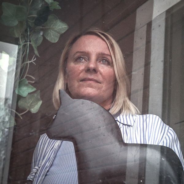 Karolin Norlén Järpeskog tittar ut genom fönstret på ett HVB-hem. Inomhusmiljö med gröna växter, en dalahäst och träpanel i bakgrunden.