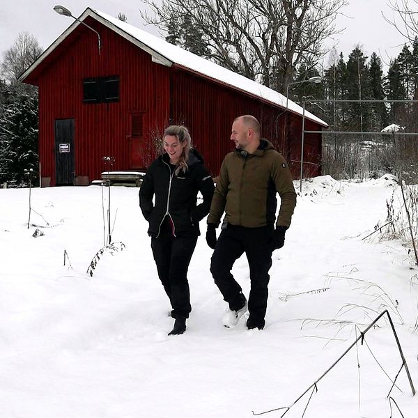 En kvinna och en man går vinterklädda längs en skogsstig med mycket snö på och runt. I bakgrunden syns en trälada.