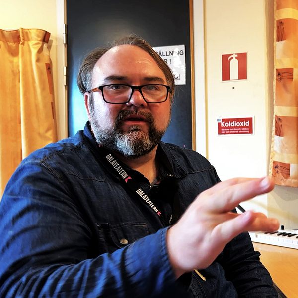 En person sitter vid ett skrivbord i en musikstudio, gestikulerande med ena handen. På skrivbordet står en laptop som visar ett musikprogram, och i bakgrunden syns ett keyboard och ljudutrustning.