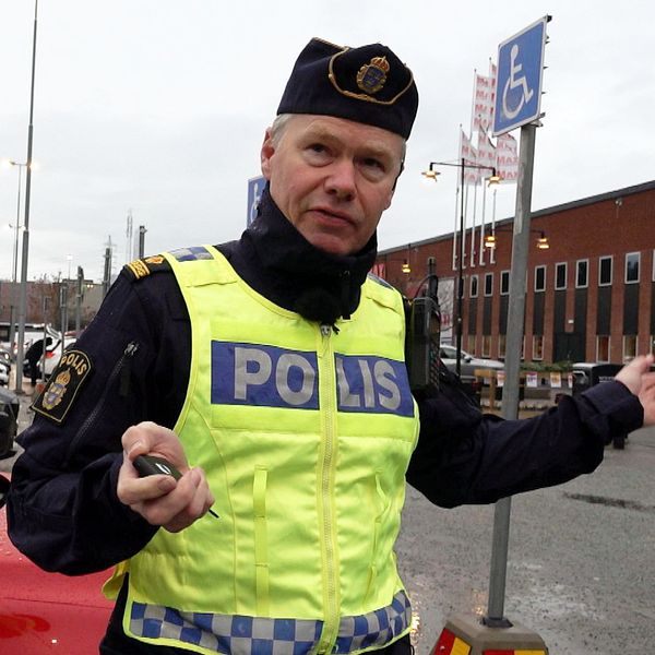 En polis står utomhus vid en parkeringsplats i Sollentuna, gestikulerande med ena handen. I bakgrunden syns en vägskylt för rullstolsanpassad parkering och en byggnad. Till höger visas en närbild av en spårsändare med kopplade kablar, märkt med en etikett.