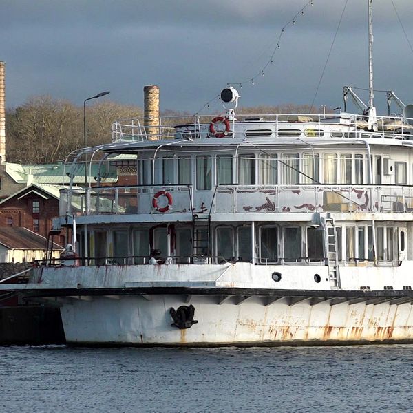 En gammal, vit passagerarbåt med rostiga detaljer ligger vid kanten av en vattenväg, omgiven av en grådaskig himmel och några byggnader i bakgrunden.