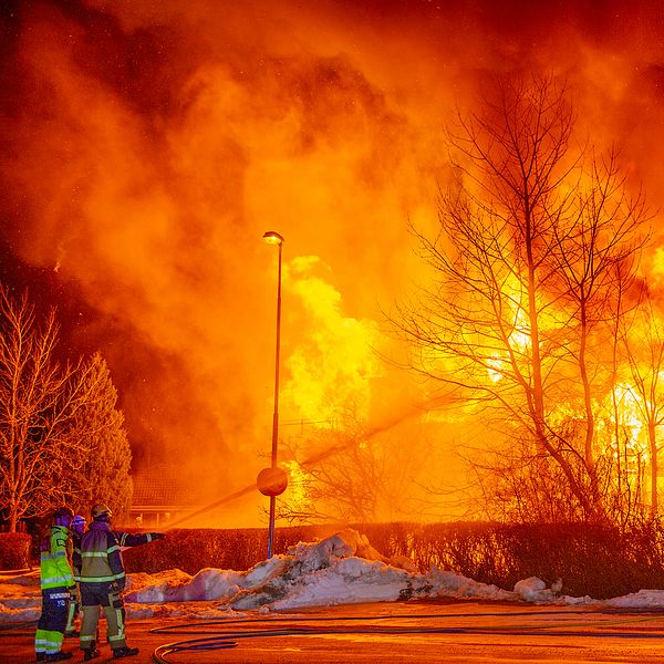 Brandmän arbetar på platsen för en massiv byggnadsbrand under natten, där intensivt orangea lågor och tjock rök stiger mot himlen. Träd och snö ses i förgrunden medan elden lyser upp omgivningen.