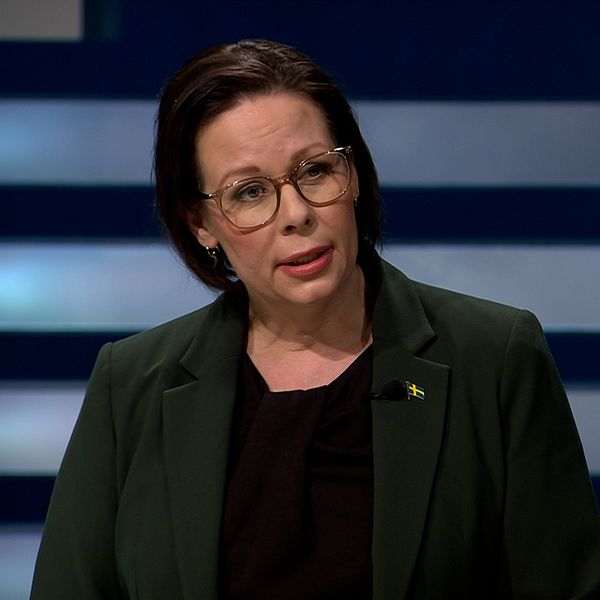 En kvinna i en grön kavaj står framför en mörk bakgrund med horisontella linjer. Hon samtalar med en annan person vid sidan av, som är delvis synlig. Kvinnan i kavajen har ett svenskt flagga-badge på sin klädd och visar en professionell attityd under diskussionen.