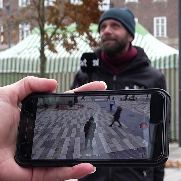 En reporter från SVT står på ett torg och filmas samtidigt av en övervakningskamera som syns i en mobiltelefon i förgrunden.