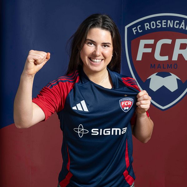 En kvinna i fotbollströja från FC Rosengård poserar med knuten näve framför sin bröst. I bakgrunden syns klubbens logotyp och färger.