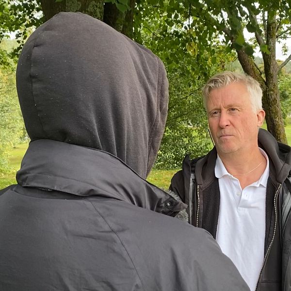 En delad bild med två delar: till vänster ses en person med mörk huva som står i en grönskande omgivning, medan den högra delen visar en röd stuga omgiven av avspärrningsband och ett träd i förgrunden under en klar himmel.