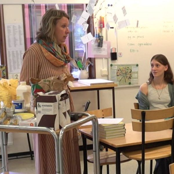 Lärare håller lektion för en grupp elever i klassrum. Elever sitter vid sina bänkar.