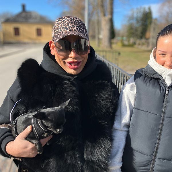 Två personer i ytterkläder utomhus, en håller en liten hund. Bakgrund med byggnad och träd.