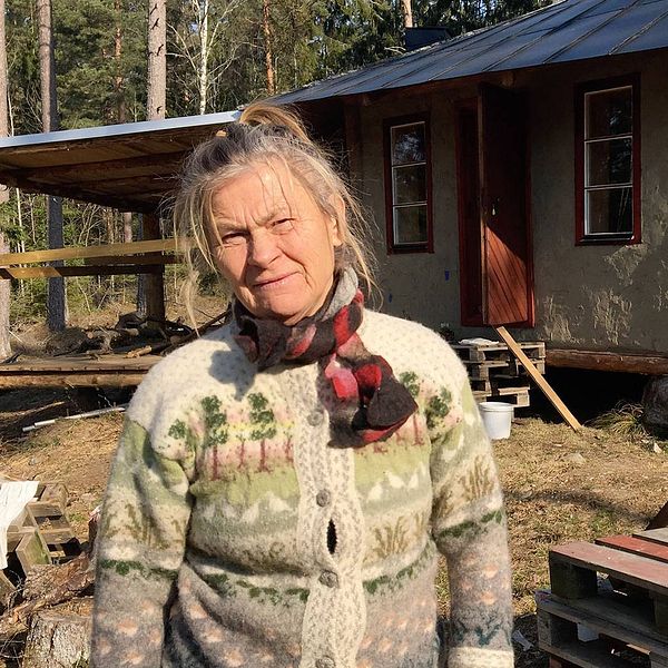 Barbro i stickad kofta står framför det lerfärgade huset med röda fönster.