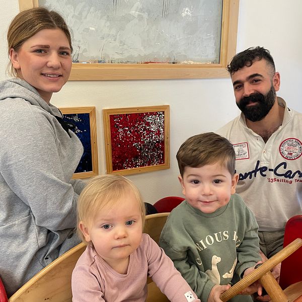 Två vuxna och två barn leker i en lekfull inomhusmiljö, med färggranna konstverk på väggen bakom dem. Till höger syns en förskolebyggnad med skylten ”Skogsborgen”, där snö ligger på marken.