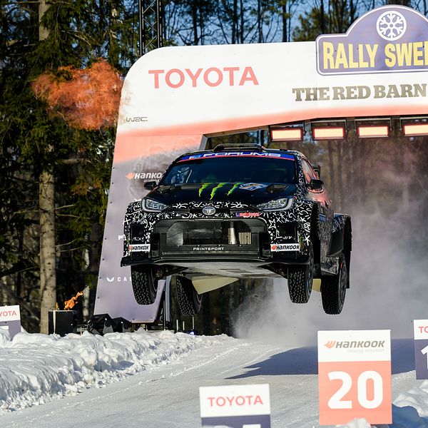 En rallybil svävar i luften efter ett hopp under Rally Sweden vid The Red Barn Arena. Omkringliggande snö och träd indikerar vinterförhållanden. Skyltar markerar sträcknummer och hastighet.