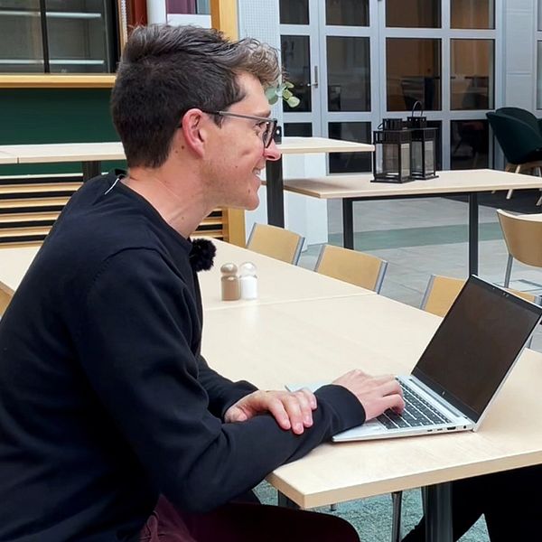 Två personer sitter vid ett långt bord i en modern inomhusmiljö. En person arbetar på en bärbar dator medan den andra håller en telefon.