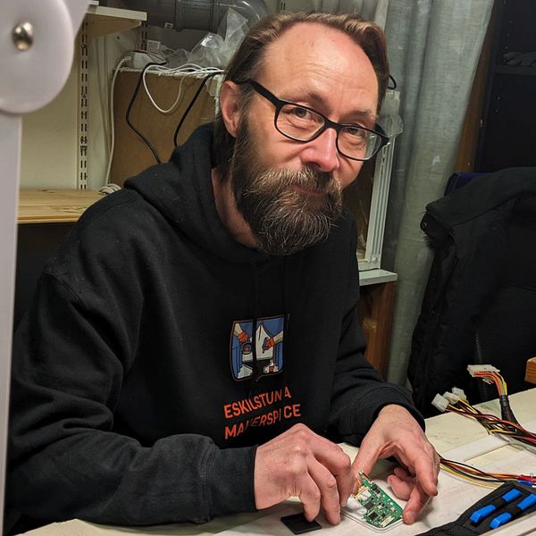 Tommie Zunko, ordförande i föreningen Eskilstuna makerspace, sitter vid ett arbetsbord fyllt av tekniska prylar och apparater och arbetar. Bredvid honom syns en figur som dansar och håller i ett huvud.