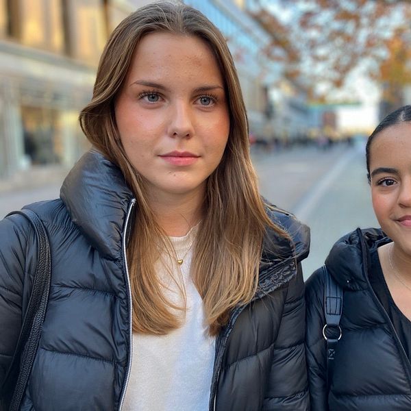 Två personer står tillsammans utomhus, iförda svarta jackor, på gågata i Uppsala med träd och byggnader i bakgrunden.