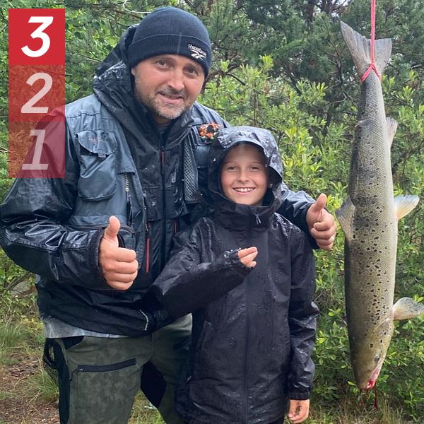 Vuxen och barn i regnkläder visar en stor upphängd fisk och ger tummen upp utomhus.