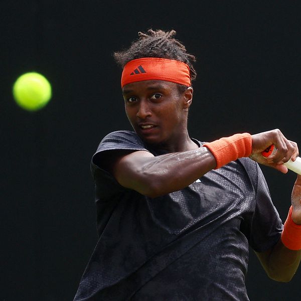 En tennisspelare i en svart tröja och med en orange svettband spelar en boll som syns i luften. Spelaren klipper en backhand med sin racket, med fokus på motståndet. Bakgrunden är mörk för att framhäva aktionen.