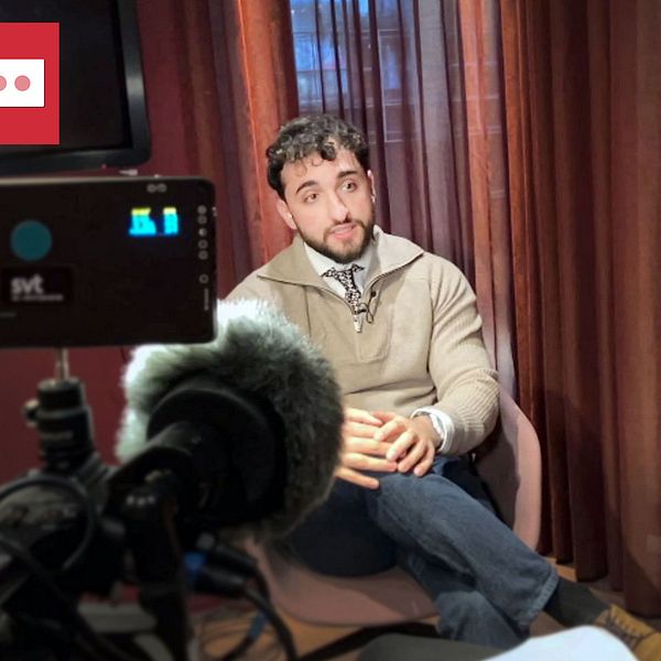 Intervjuperson i fåtölj pratar med journalist i ett rum med mörka gardiner, filmutrustning synlig.