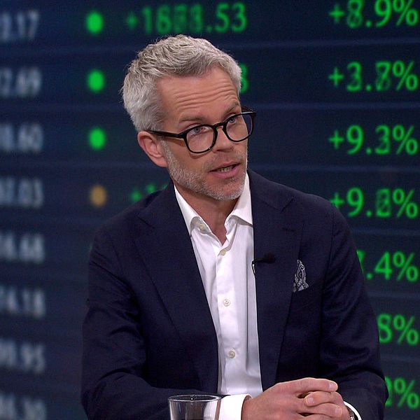 Person i kostym i tv-studio med börskurser och siffror synliga i bakgrunden.