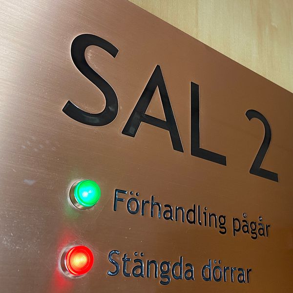 En skylt utanför en tingsrättssal i Nyköping. På skylten står det ”Sal 2”. Under det står det ”Förhandling pågår”, där det lyser en grön lampa bredvid. Under det står det ”Stängda dörrar” där det lyser en röd lampa bredvid.