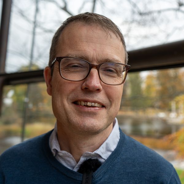 Regionstyrelsens ordförande Patrik Stenvard (M) står i fokus med bakgrund av en grön och orange höstlandskap genom ett stort fönster. Ljuset är mjukt och naturen syns tydligt, vilket skapar en lugn atmosfär.