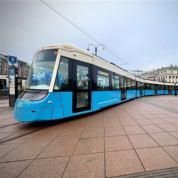 En modern spårvagn i ljusblå färg står på en torgyta med stora plattor. I bakgrunden syns historisk arkitektur och några personer som går förbi, med en lätt dimma över området.