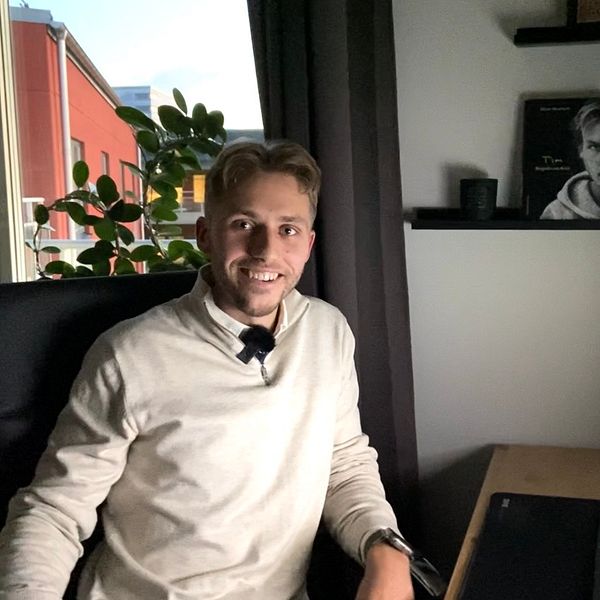 Oliver Berg sitter vid ett skrivbord i ett kontorsutrymme, klädd i en ljus tröja och skjorta. På skrivbordet finns en dator med en färgglad skärm som visar en graf eller diagram, och i bakgrunden syns en växt och hyllor med böcker eller dekorationer. Rummet har fönster som släpper in ett svagt ljus.