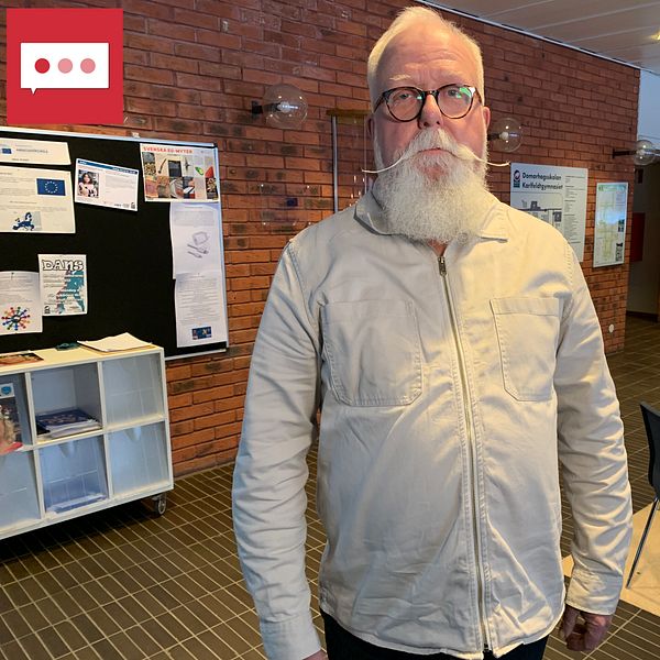 En man med lång spetsig mustasch tittar in i kameran i en skolcafeteria.
