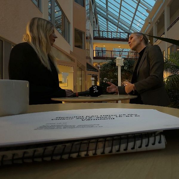 Två personer står och samtalar i en ljus, växtfylld atrium, med en stor glasad takstruktur. I förgrunden syns en anteckningsbok och en mugg, vilket tyder på att de diskuterar ett arbetsrelaterat ämne.