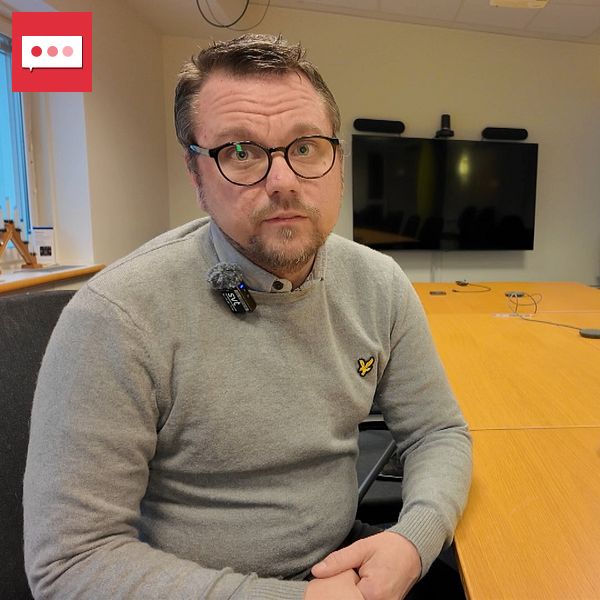 En person sitter vid ett konferensbord i ett rum med modern inredning. Framför honom står en laptop och på bordet syns mikrofon och mötesutrustning. I bakgrunden finns en stor skärm. Rummet är ljust och har stora fönster som ger insyn till utomhusmiljön.