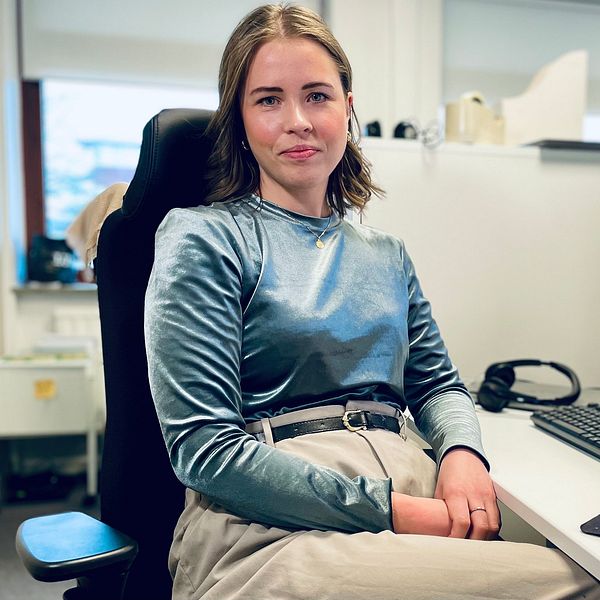 En person sitter vid en arbetsstation med en dator framför sig. Bakgrunden visar en kontorsmiljö med ljusa fönster. Datorn visar en webbplats, och det finns en mus och ett tangentbord på skrivbordet. Personen bär en långärmad tröja och byxor, och sitter avslappnat i en ergonomisk stol.