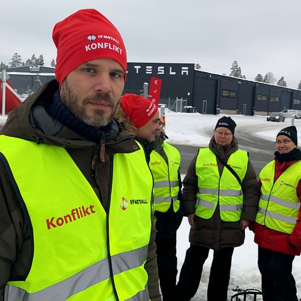 Flera personer med gula västar där det står ”konflikt” i röd text står framför Teslas svarta anläggning.