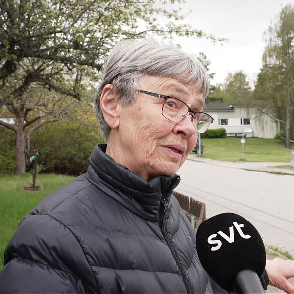 Reporter håller SVT-mikrofon mot en äldre kvinna, polis står bakom avspärrning på lugn villagata.