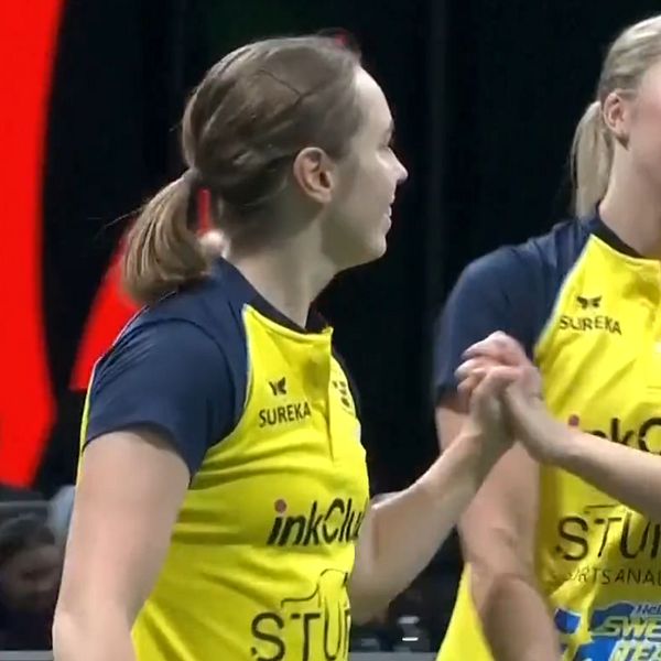 Två kvinnliga spelare i sportkläder med gul och blå design, står på en sportarena och interagerar med varandra under en paus eller ett vinstmoment. De verkar glada och uppmuntrande mot varandra. Bakgrunden visar publiken och en ljusinstallation som skapar en sportig atmosfär.