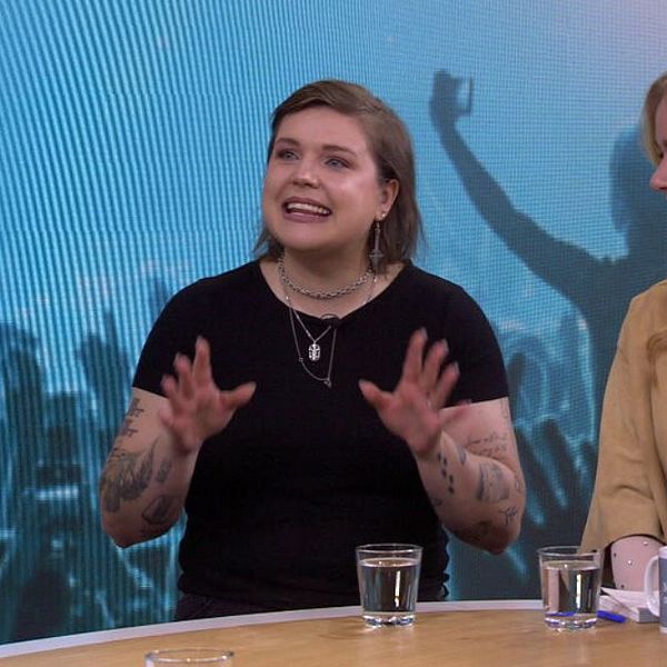 Två personer sitter vid ett bord i en tv-studio, bakom dem skymtar en konsertpublik.