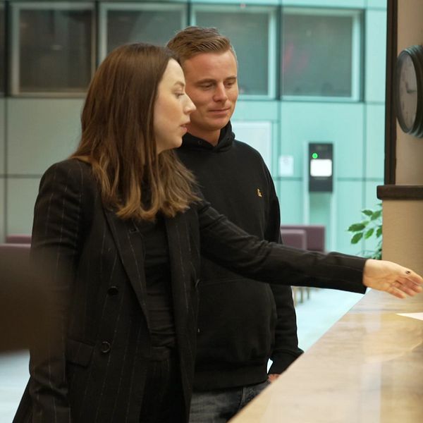 Jonathan Ravelin och Elsa Häggård står vid en receptionsdisk och lämnar över ett dokument. Bakgrunden visar en modern kontorsmiljö med växter och en klocka på väggen. Skärmen på disken är delvis synlig.