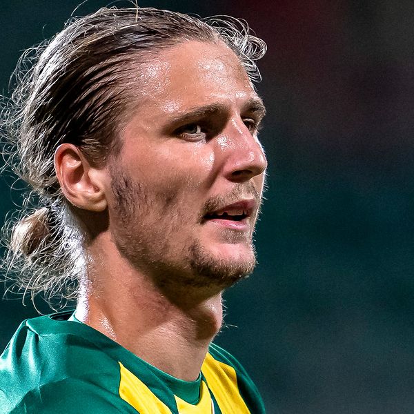 En delad bild som visar två fotbollsspelare. Till vänster, en spelare i en grön och gul tröja med kort hår sett i profil. Till höger, en annan spelare iförd en orange väst som håller en fotboll, stående på en plan omgiven av träd i bakgrunden.