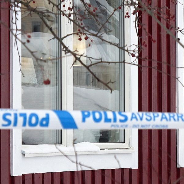 Fönster på ett rödbrunt hus inhägnat med blåvita polisavspärrningsband och grenar från ett träd i förgrunden.