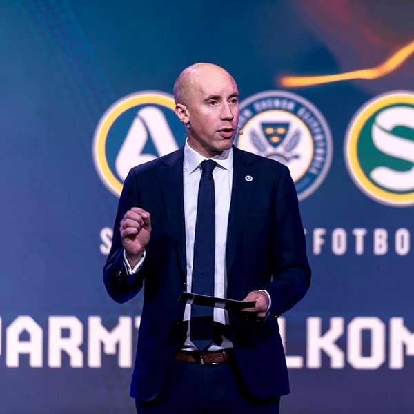 Man i kostym på scen. Banners med fotbollssymboler och text ”Varmt välkommen!” i bakgrunden.